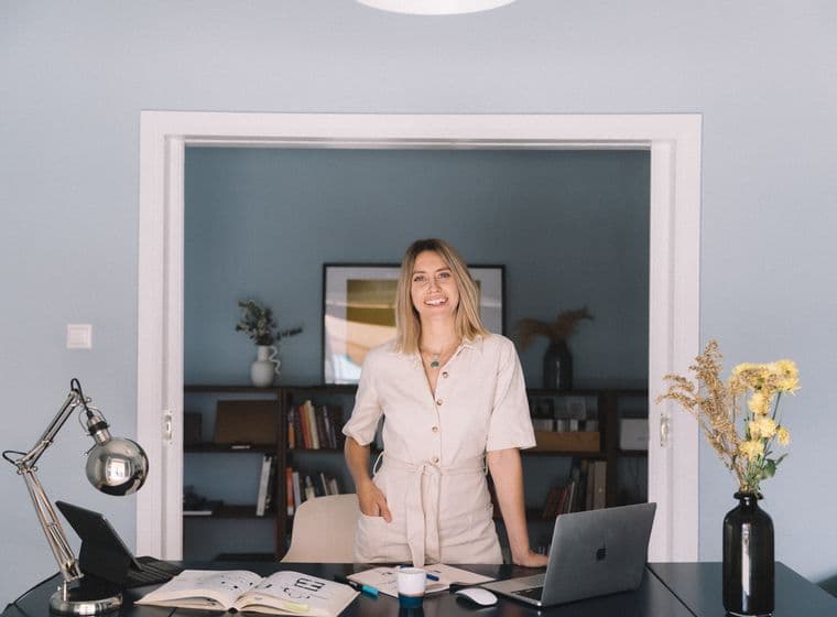 Μια χαμογελαστή γυναίκα με μπεζ φόρμα στέκεται σε ένα γραφείο με ένα λάπτοπ και λουλούδια σε ένα μπλε δωμάτιο.
