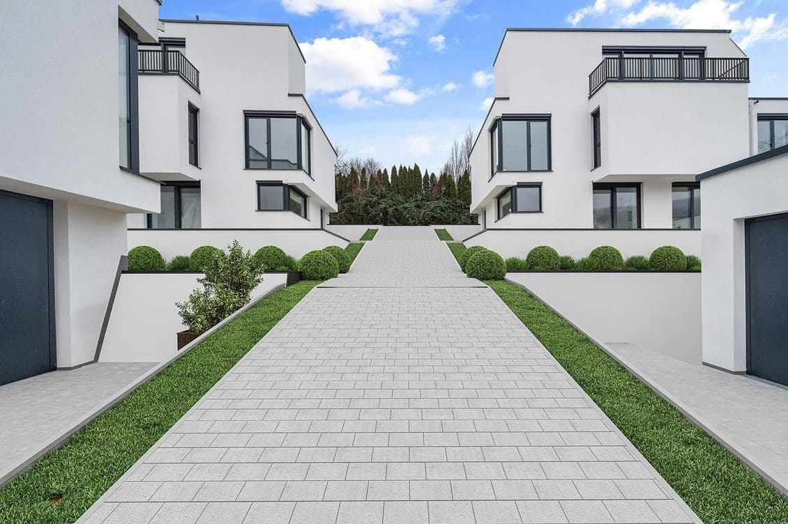 Moderne weiße Gebäude mit großen Fenstern flankieren einen breiten gepflasterten Weg, der von grünem Gras und Büschen gesäumt ist, unter einem teilweise bewölkten Himmel.