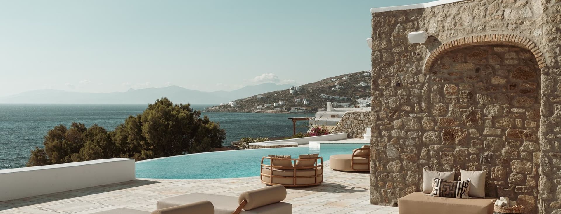 Poolside terrace with beige loungers beside a stone wall, an infinity pool, and a sea view over a hillside village.