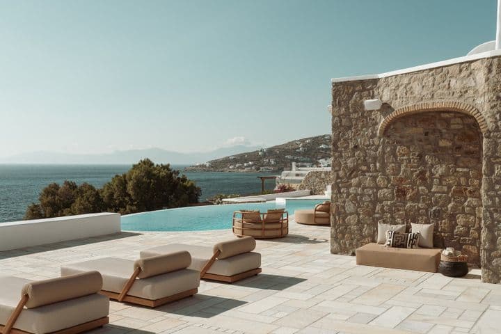 Poolside terrace with beige loungers beside a stone wall, an infinity pool, and a sea view over a hillside village.