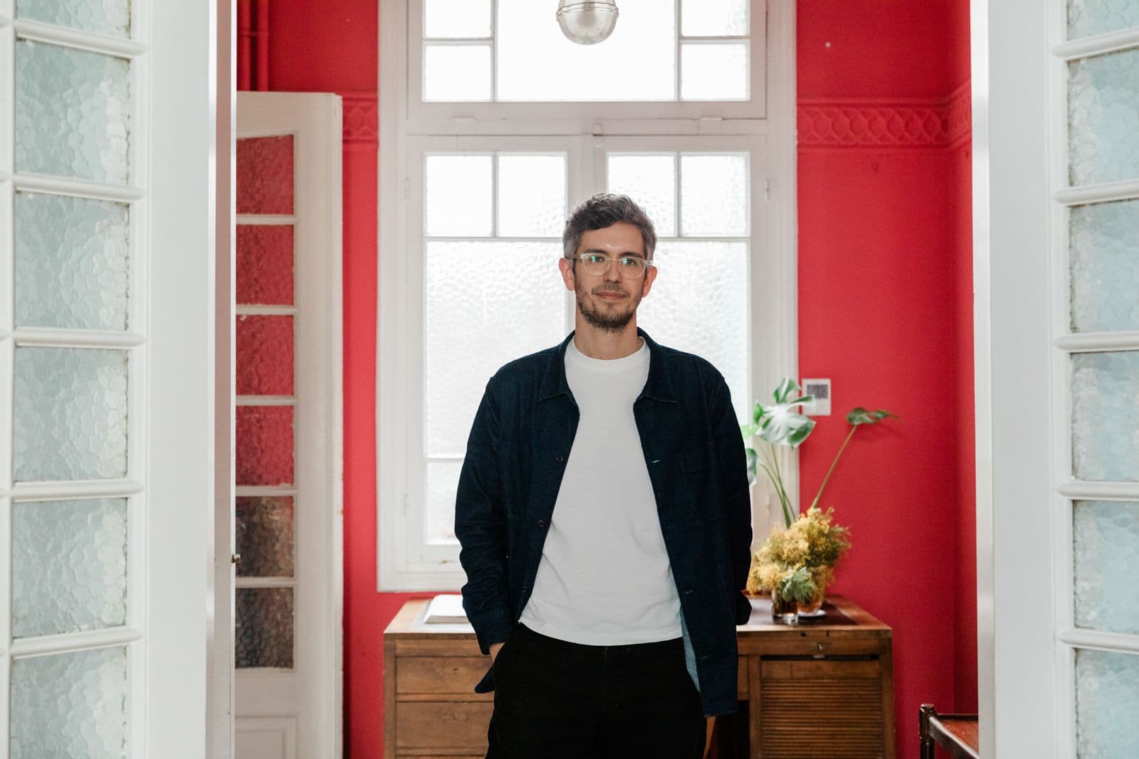 Ένας άντρας με γυαλιά στέκεται στην πόρτα ενός κόκκινου δωματίου με ένα άσπρο παράθυρο και μια ξύλινη ντουλάπα.