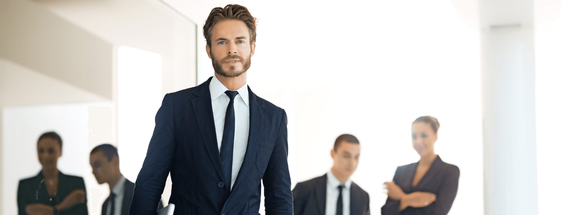 Un home amb vestit camina cap a la càmera, sostenint un portàtil, amb companys de feina al fons.