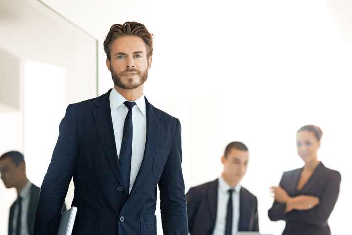 Un home amb vestit camina cap a la càmera, sostenint un portàtil, amb companys de feina al fons.