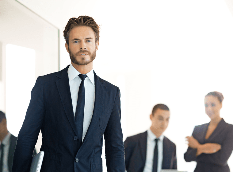 Un home amb vestit camina cap a la càmera, sostenint un portàtil, amb companys de feina al fons.