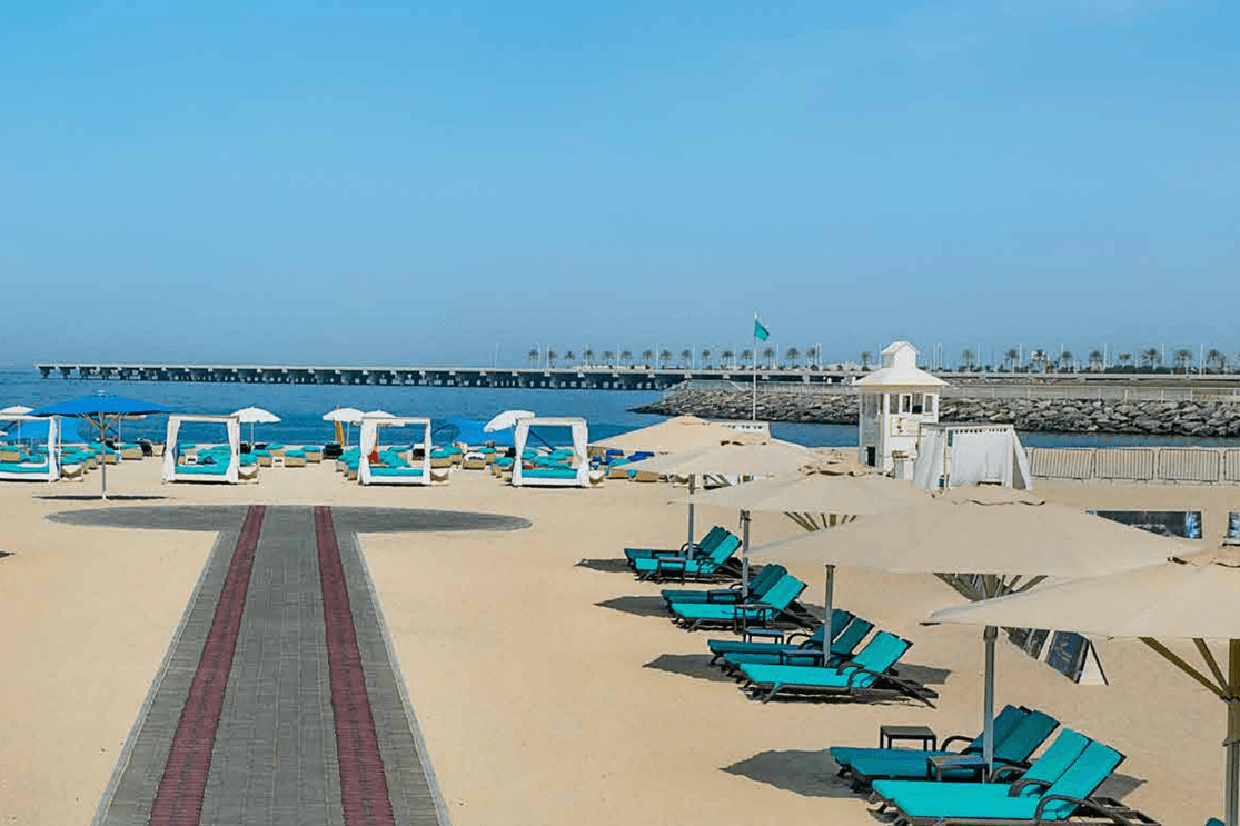 A beach view with empty loungers and umbrellas on sand, a pathway leading to the sea, and a distant pier under a clear blue sky.