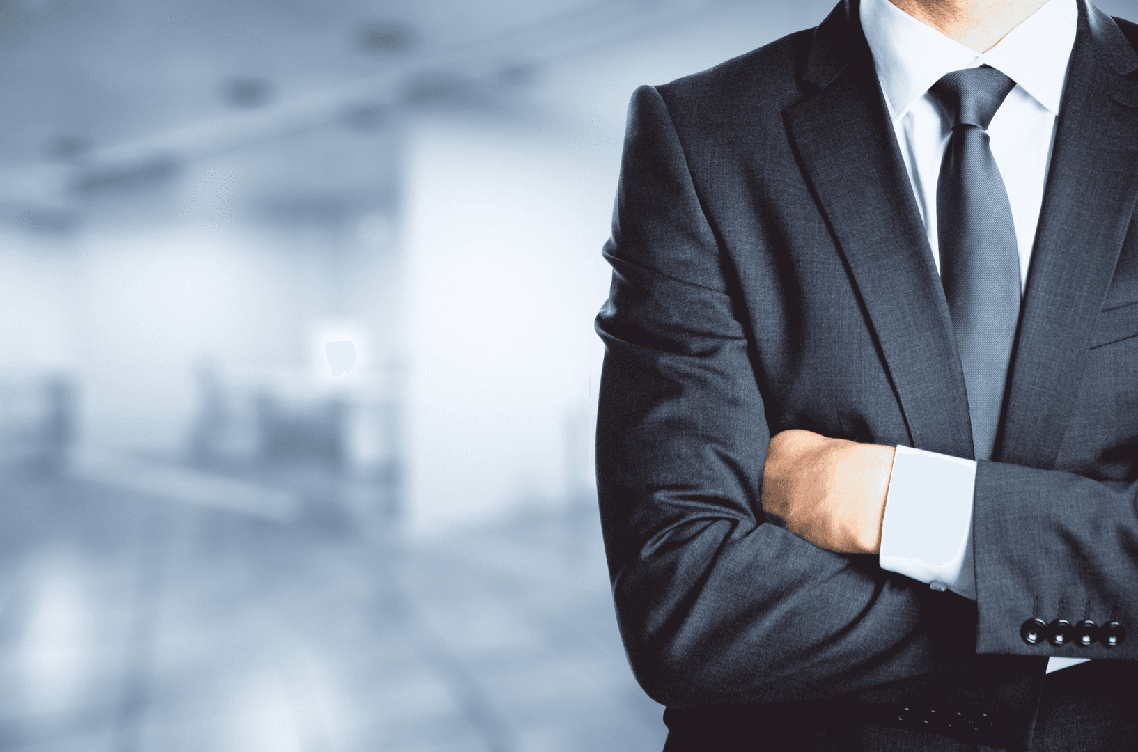 A man in a dark gray suit and tie stands with his arms crossed in front of a blurred office background.