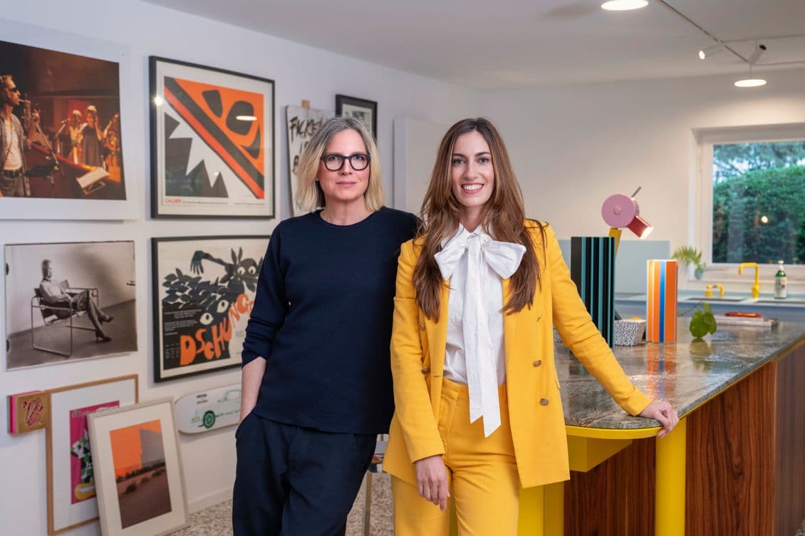 Two women stand in a modern, art-filled room. One wears a black outfit, the other a yellow suit with a white bow blouse. Framed art decorates the walls.