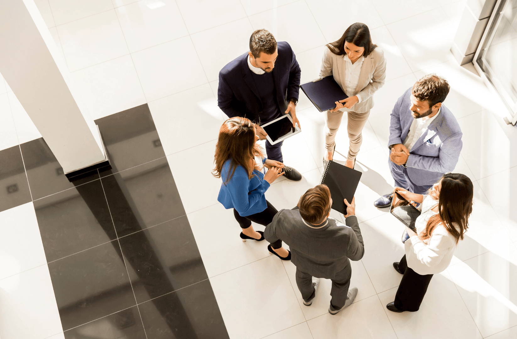 Overhead shot of six business people in suits standing in a circle, holding tablets and notebooks. The floor is tiled in black and white.