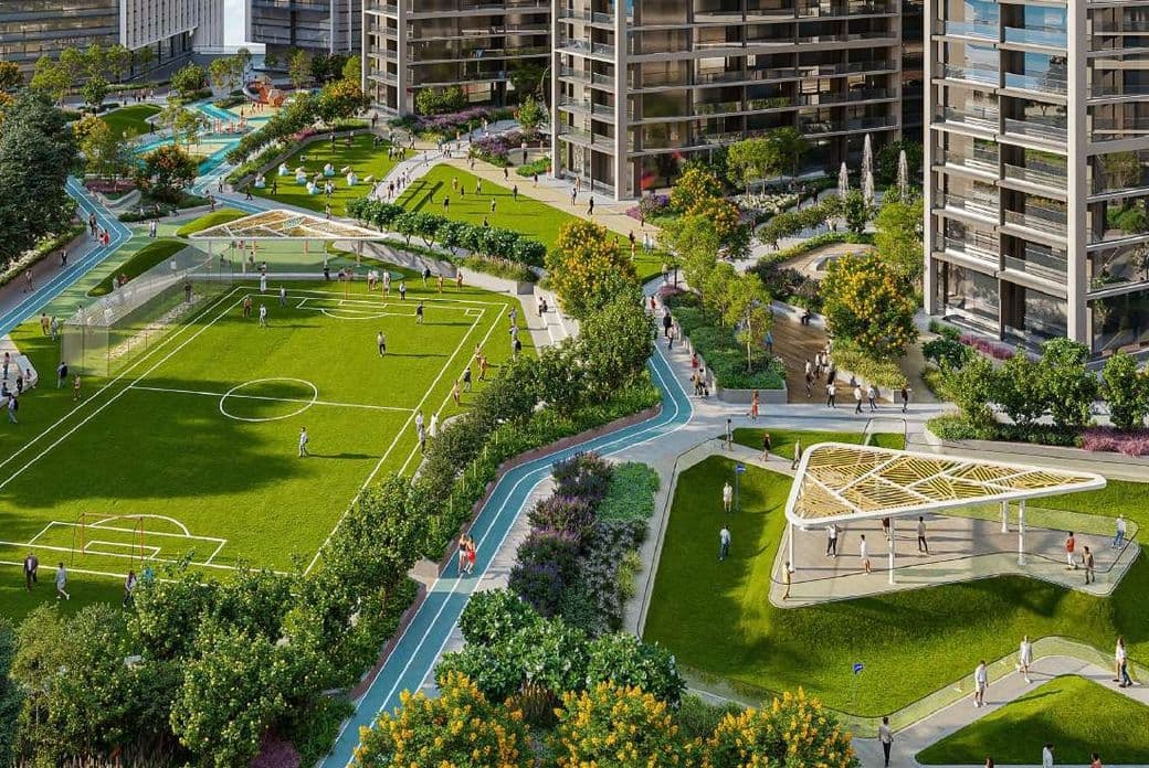 Aerial view of a modern residential complex with landscaped gardens, a soccer field, walking paths, and recreational areas surrounded by tall buildings.