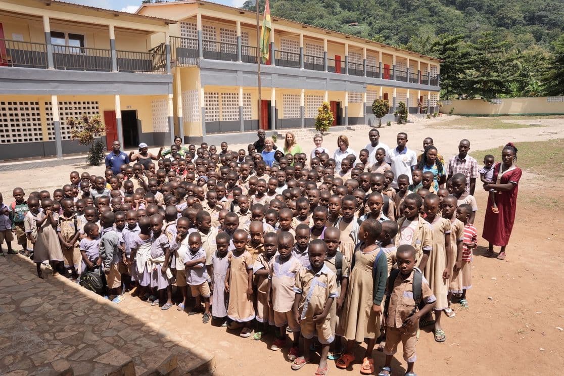 A large group of children in school uniforms and adults stand in front of a two-story school building with a mountainous backdrop.