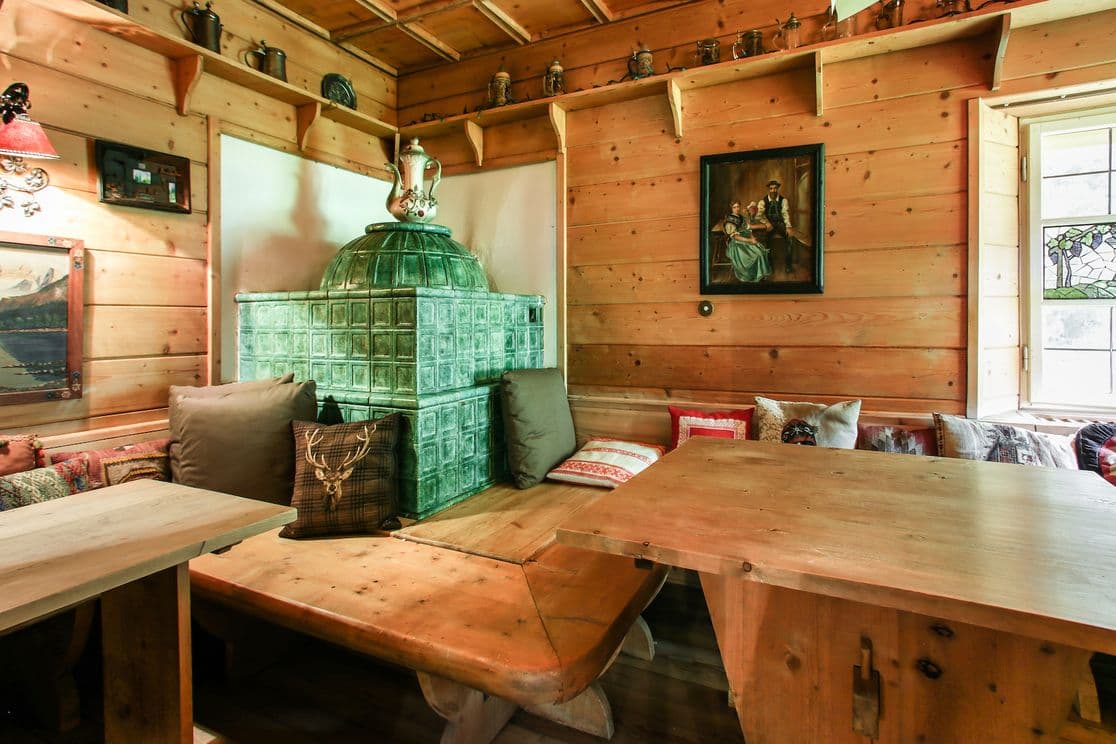 Gemütliche Holzstube mit grünem Kachelofen, Holzbänken, Kissen und einem Fenster mit Buntglas. Rustikale Einrichtung und warme Beleuchtung.