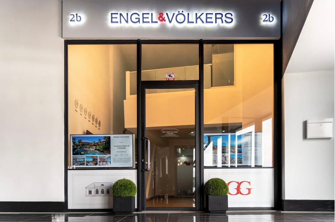Front entrance of Engel & Völkers office in Mauritius with glass doors, illuminated sign, digital displays, and decorative plants.