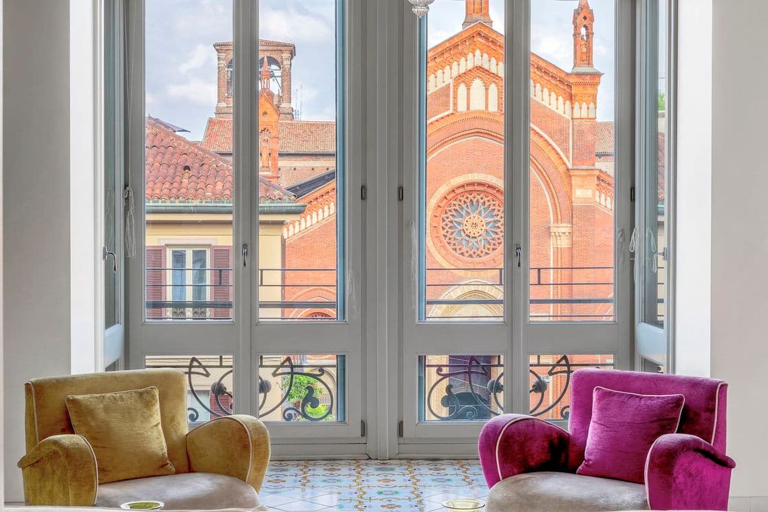 Zwei bunte Sessel stehen vor großen Fenstern mit Blick auf ein historisches Backsteingebäude mit kunstvoller Architektur und einem runden Buntglasfenster.