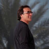 A smiling man in glasses and a black shirt stands in front of a palm tree backdrop.
