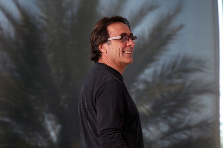 Ein lächelnder Mann mit Brille und schwarzem Hemd steht vor einer Palme.