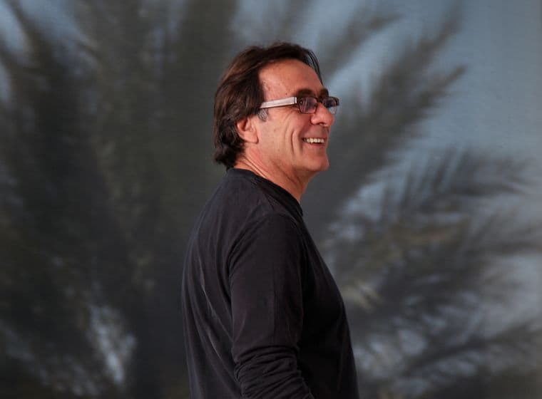 Ein lächelnder Mann mit Brille und schwarzem Hemd steht vor einer Palme.