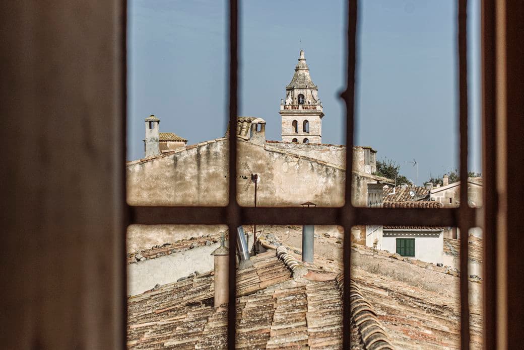 Vista a través d'una finestra de fusta que mostra els teulats i una torre de campanar històrica a Sencelles, Mallorca.
