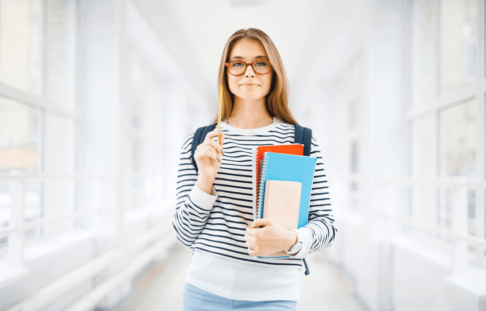A young woman with glasses holds books and a pencil in a bright hallway.