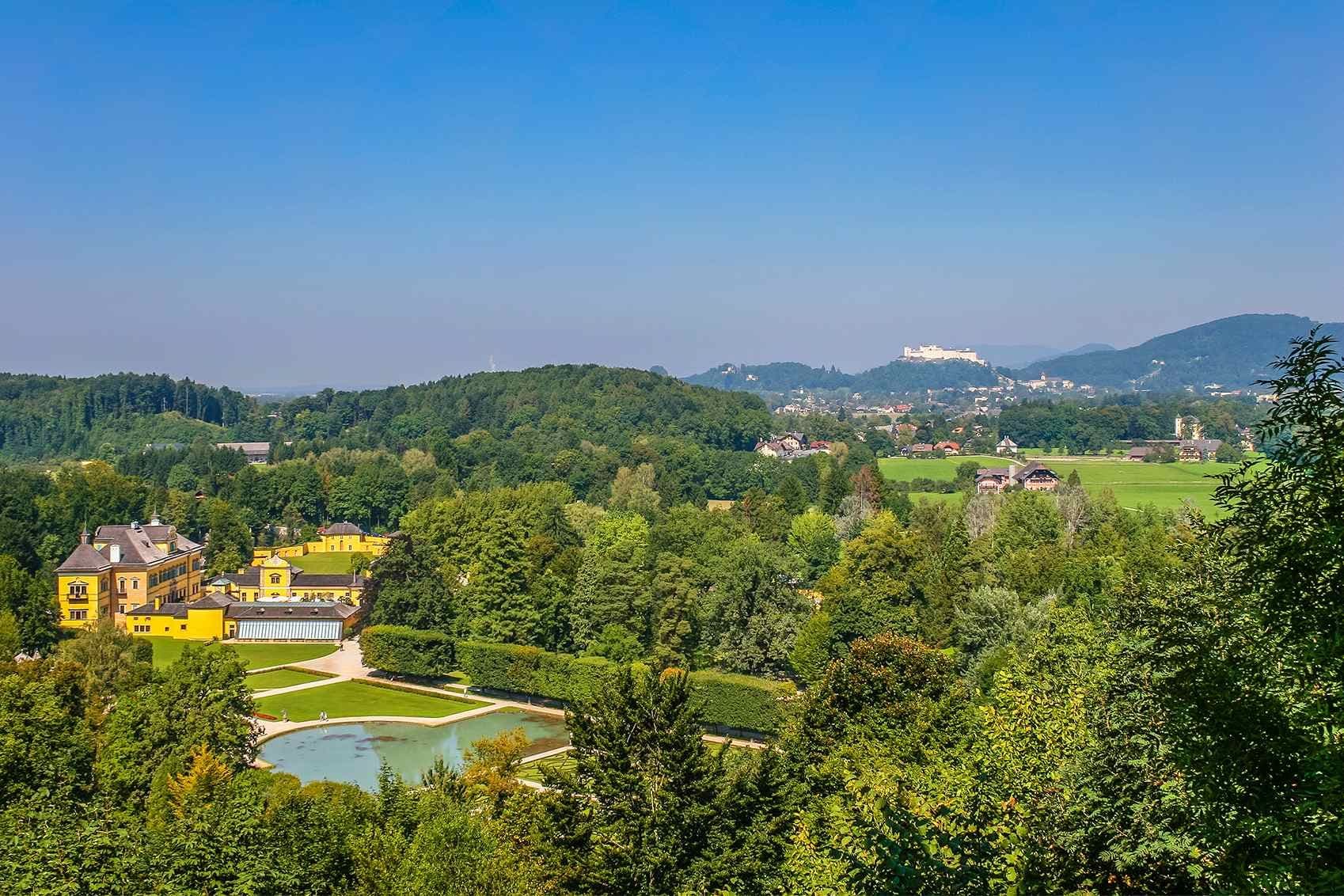 Blick auf Schloss Leopoldskron mit gelbem Barockgebäude, gepflegtem Garten und Teich, im Hintergrund die Festung Hohensalzburg und grüne Hügellandschaft.