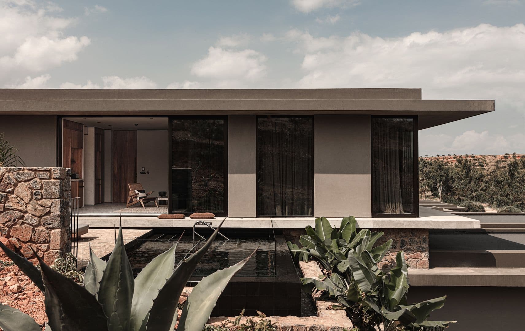Μοντέρνο εξωτερικό σπιτιού με επίπεδη στέγη, πέτρινους τοίχους και μια σκούρα πισίνα. Μεγάλες γυάλινες πόρτες αντανακλούν το τοπίο. Πράσινα φυτά στο προσκήνιο.