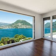 Habitación luminosa con pisos de madera y una gran ventana con vistas a un lago azul y montañas.