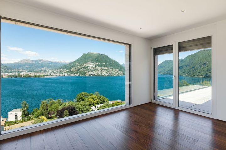 Habitación luminosa con pisos de madera y una gran ventana con vistas a un lago azul y montañas.
