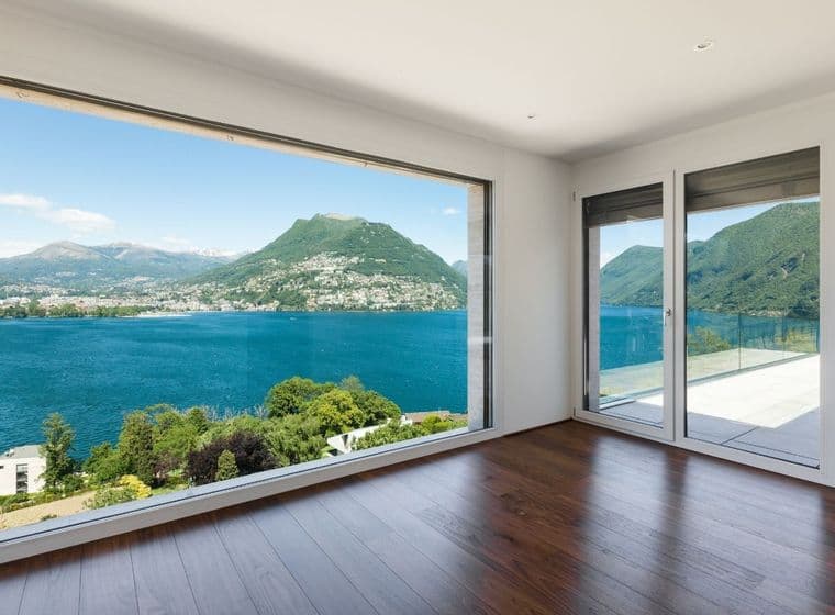 Habitació lluminosa amb terres de fusta i una gran finestra amb vista a un llac blau i muntanyes.