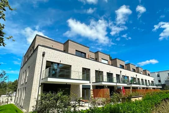 Eine Reihe moderner Backsteinreihenhäuser mit Glasbalkonen unter strahlend blauem Himmel, umgeben von grünen Sträuchern.
