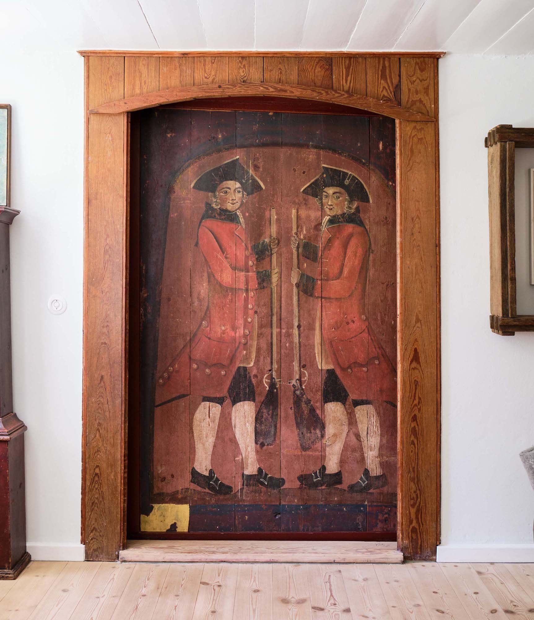 Large wooden panel with painted image of two soldiers in red coats, holding rifles, set in a wooden frame against a white wall.