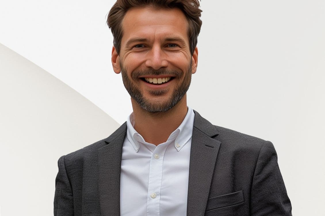 Homem sorrindo vestindo terno cinza e camisa branca contra um fundo branco. Ele tem cabelo castanho e barba.