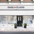 Modern Engel & Völkers real estate office storefront with white classical architecture featuring columns and decorative moldings. The brand name 'ENGEL&VÖLKERS' is displayed in large black letters with a red ampersand above the entrance. The shopfront has large windows displaying property listings in illuminated display cases on both sides of the central glass door entrance. Two decorative planters with green topiary bushes flank the doorway. The building sits on a cobblestone street with traditional European architecture.
