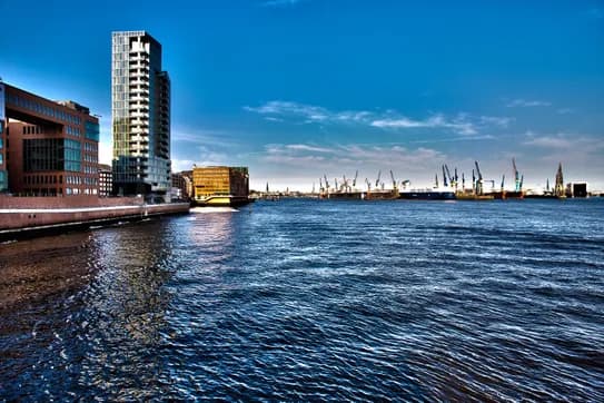 Blick auf den Hafen mit einem hohen Glaswolkenkratzer und Backsteingebäuden auf der linken Seite, Kränen und Schiffen auf der anderen Seite des Wassers unter blauem Himmel.