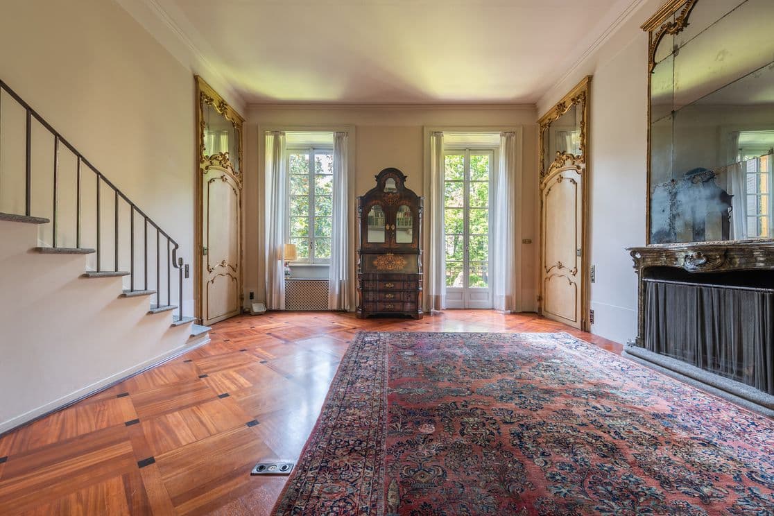 Eleganter Raum mit verzierten Spiegeln, Vintage-Möbeln, großen Fenstern und einem gemusterten Teppich auf poliertem Holzboden. Treppe auf der linken Seite.