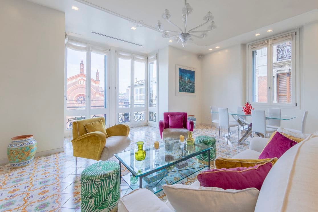 Helles Wohnzimmer mit buntem Fliesenboden, modernen Möbeln, Couchtisch aus Glas und großen Fenstern mit Blick auf ein Gebäude mit Bogenfenstern.