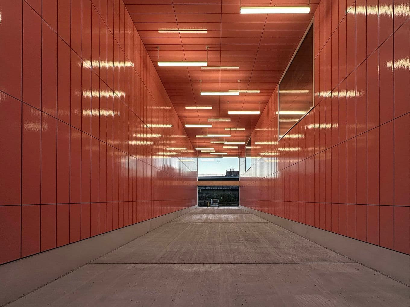 A corridor with bright orange walls and ceiling illuminated by linear lights, leading to an outdoor view in the distance.