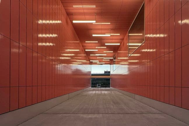 A corridor with bright orange walls and ceiling illuminated by linear lights, leading to an outdoor view in the distance.