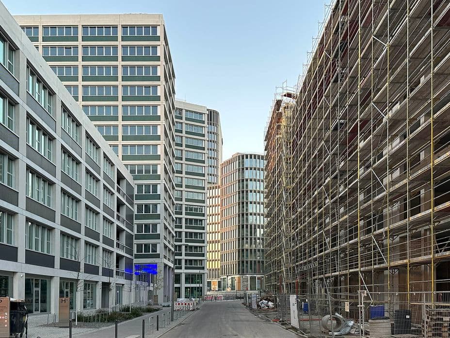 A narrow street flanked by modern office buildings under construction with scaffolding, leading to a cluster of tall, glassy structures.