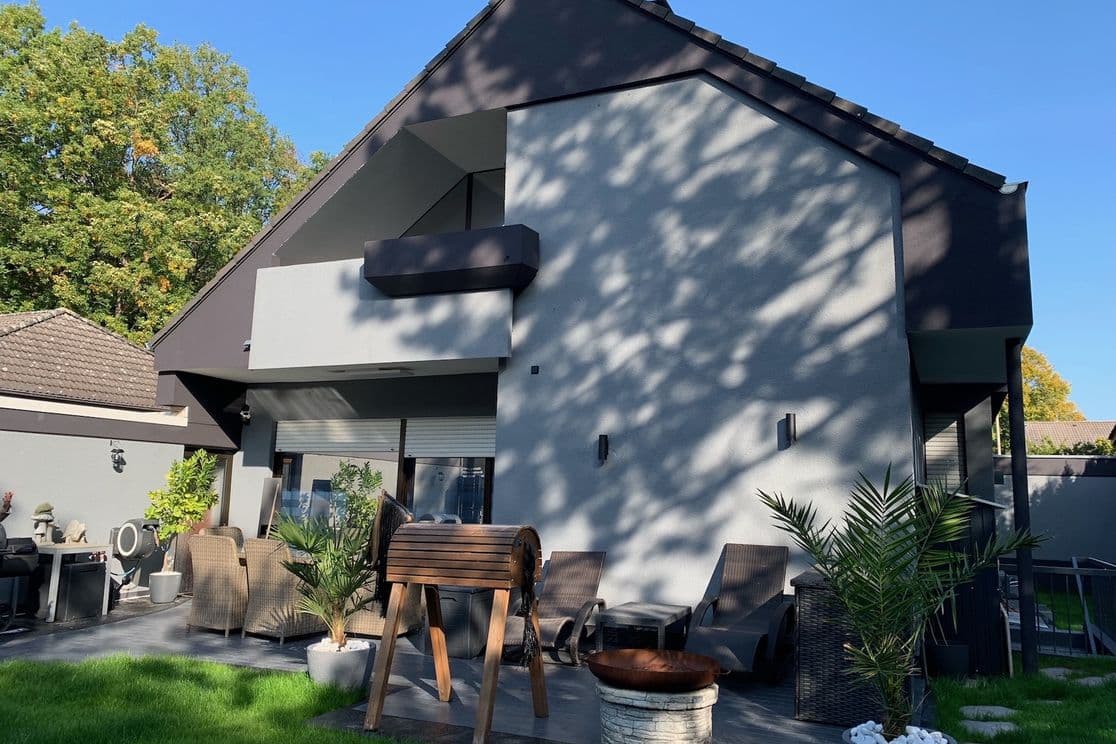Modern two-story house with a patio, wicker furniture, potted plants, and a lush green lawn under a clear blue sky.