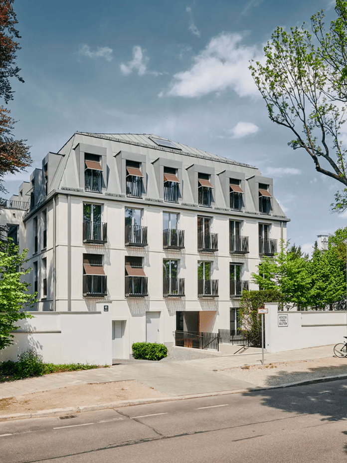 Vierstöckiges modernes Wohnhaus mit symmetrischen Fenstern, umgeben von Grünflächen und Bäumen, unter einem strahlend blauen Himmel in einer ruhigen Straße.