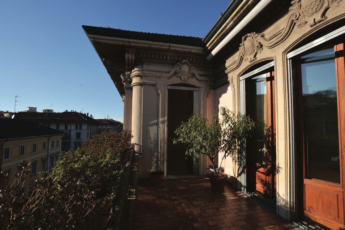 Ein sonnenbeschienener Balkon mit kunstvoller Architektur, Topfpflanzen und Blick auf die Nachbargebäude unter einem klaren blauen Himmel.