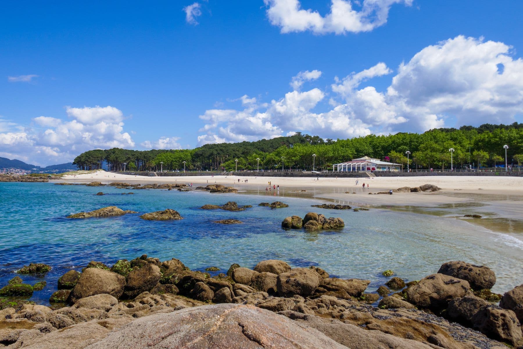 Playa de Samil in Vigo, Spanien. Zusehen sind Felsen, das Meer, Bäume und ein Restaurant.