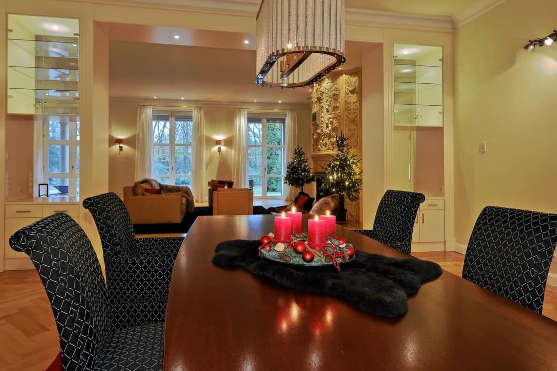 Elegant dining room with dark wooden table, Advent arrangement with three burning candles on top, and view of a Christmas-decorated living room in the background.