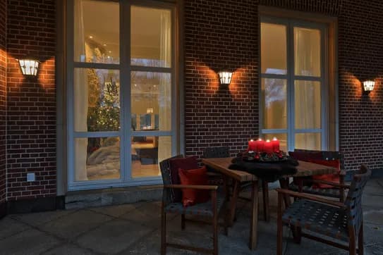 Evening scene of a terrace with a brick wall, bright wall lamps and a wooden table with an Advent arrangement with burning candles. An illuminated Christmas tree is visible inside through the large windows.