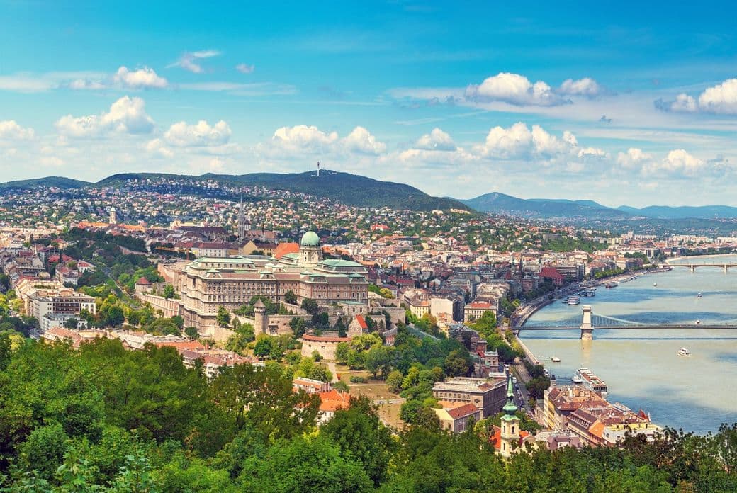 Panoramic view of Budapest with Buda Castle, the Danube River, and the Chain Bridge under a blue sky with scattered clouds.