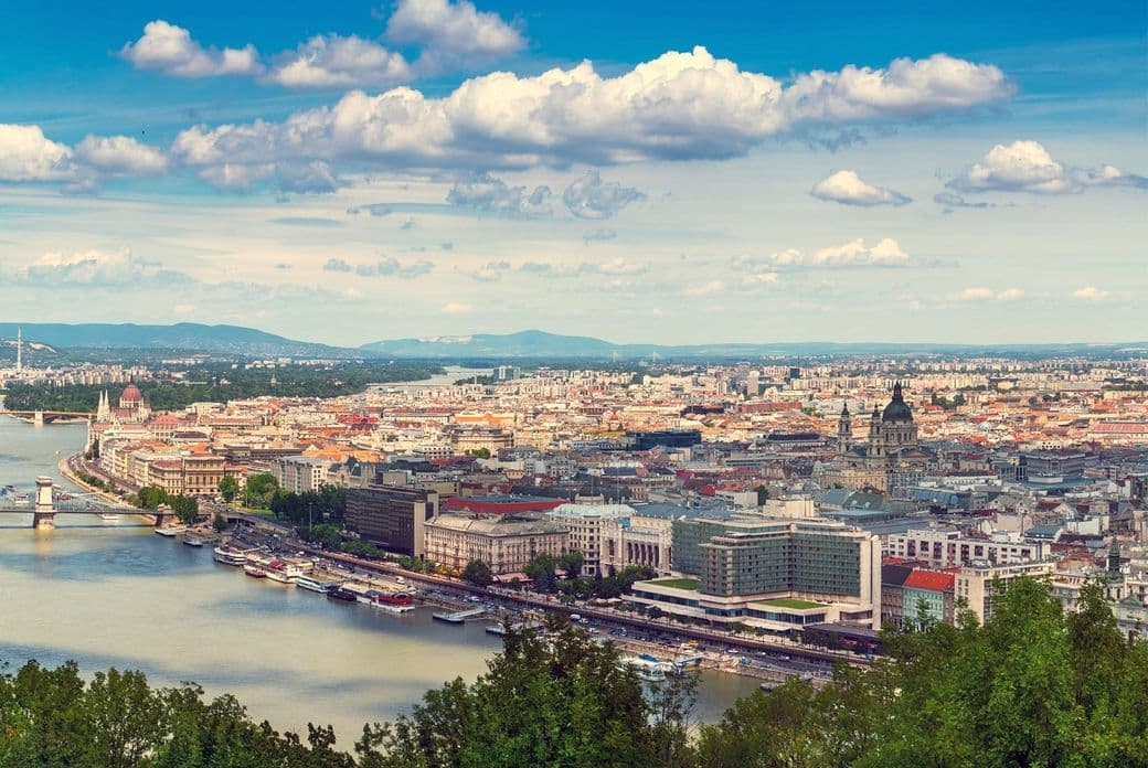 Aerial view of Pest with the Danube River, historic buildings, and bridges under a partly cloudy sky.