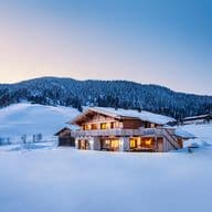 A cozy wooden chalet with glowing windows surrounded by snowy hills and forest under a clear evening sky.
