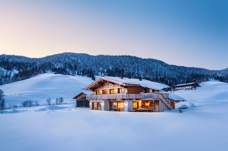 A cozy wooden chalet with glowing windows surrounded by snowy hills and forest under a clear evening sky.