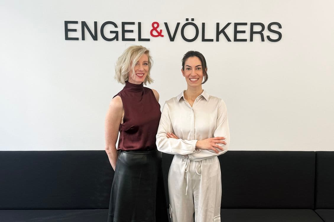 Two women standing in front of an Engel & Völkers sign, smiling. One wears a burgundy top, the other a white outfit, both appear professional.