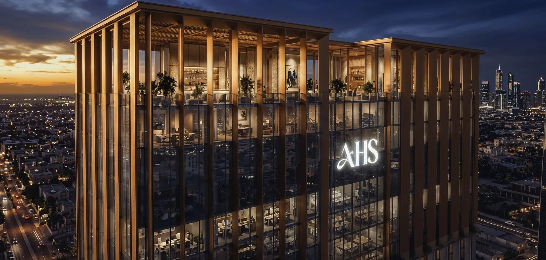 Evening view of a glass skyscraper with gold vertical frames; warm interiors glow; bright AHS logo on the facade; distant city skyline.