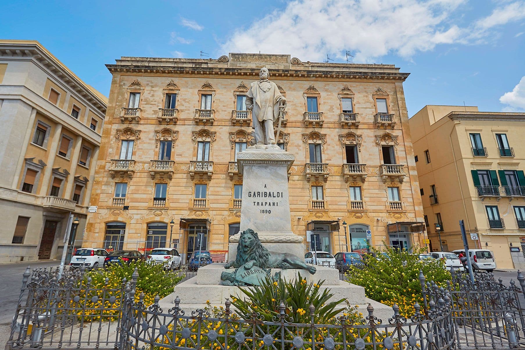 Statue of Garibaldi in Trapani, Italy, set in a fenced garden with flowers, in front of a historic building under a partly cloudy sky.